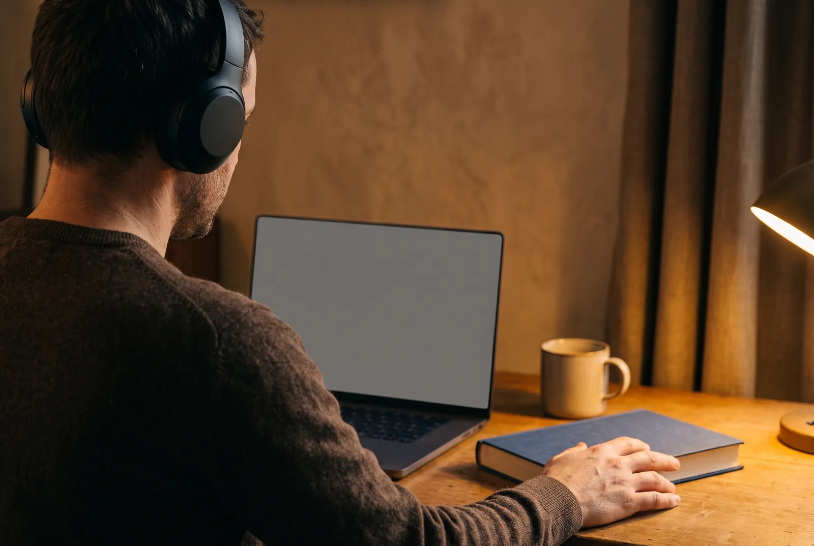A man at a desk in the evening wearing headphones during an online class, seen from behind, a closed book beside the laptop.