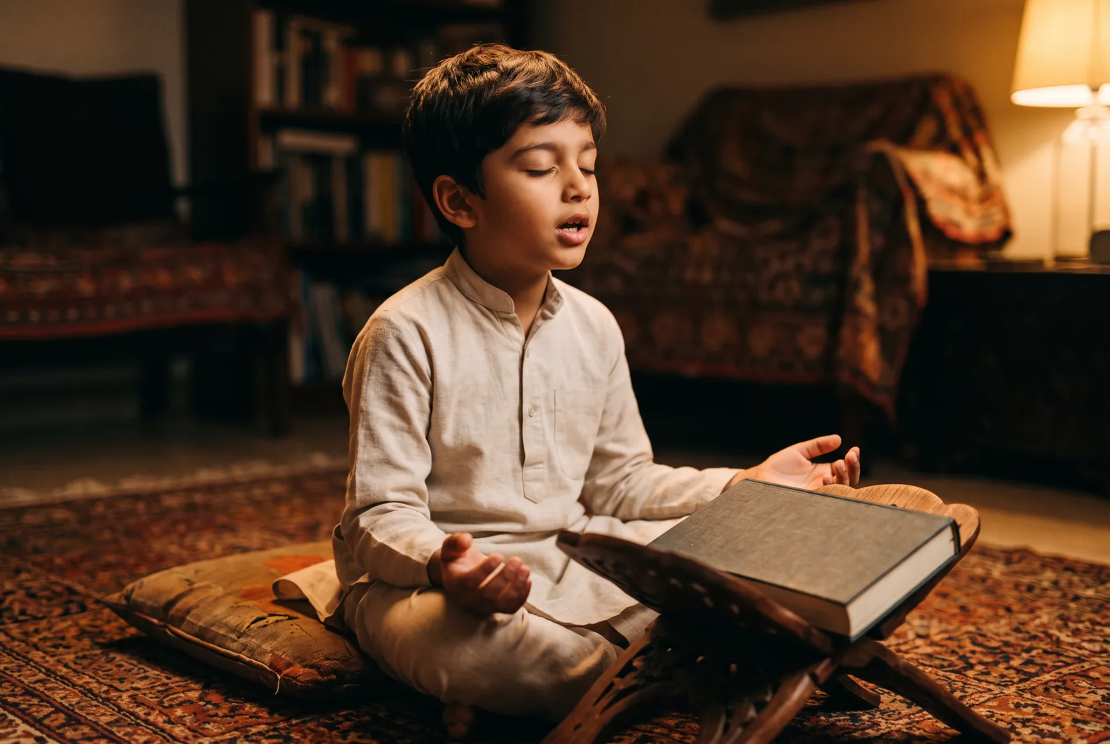 A boy reciting aloud from an open mushaf on a wooden rehal while his mother listens beside him.