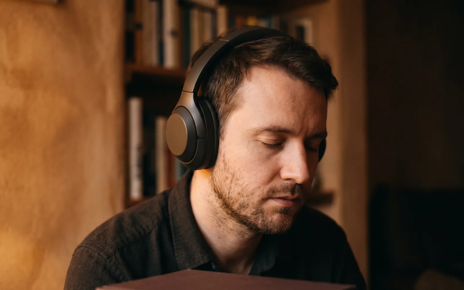 An adult wearing over-ear headphones with his eyes closed, listening closely, in a warm room.