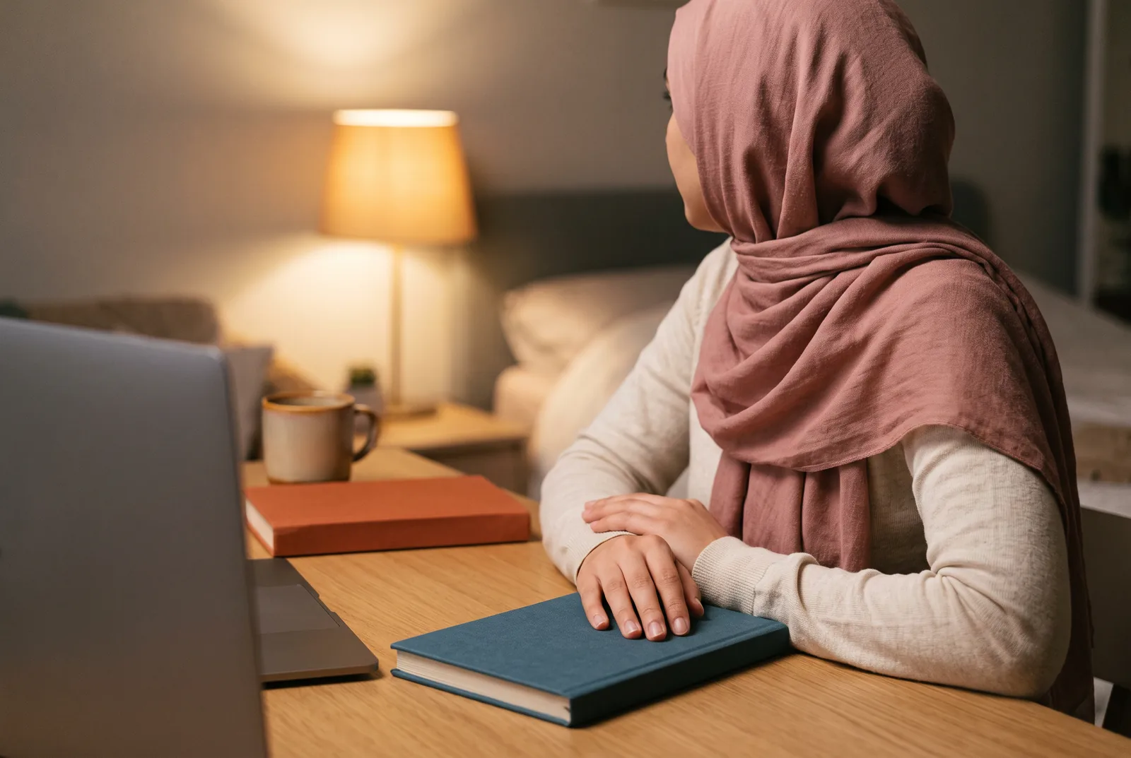 A woman in a headscarf at a table in the evening during an online class, a closed book under her hand.